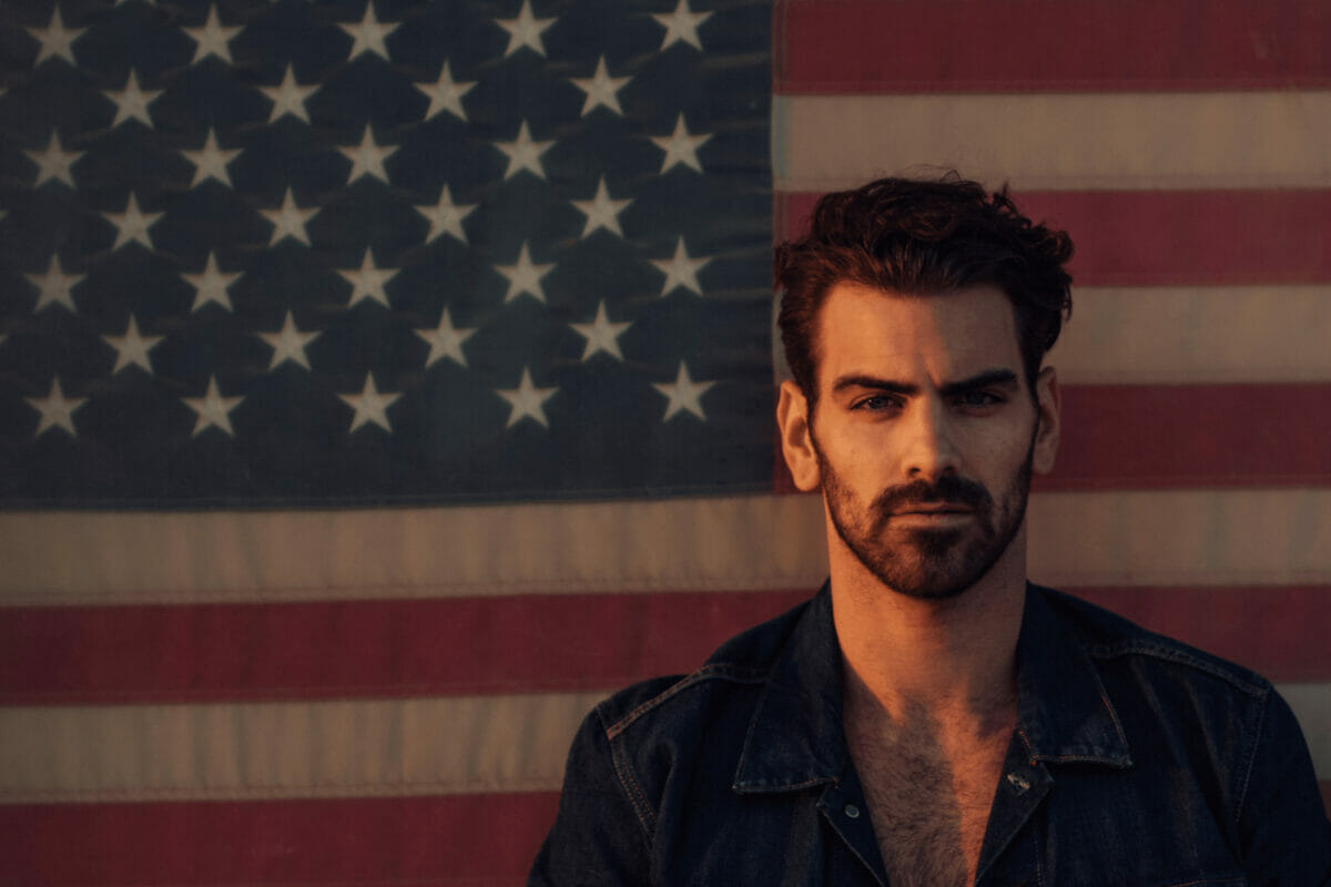 Nyle DiMarco with a rugged look in front of an American flag